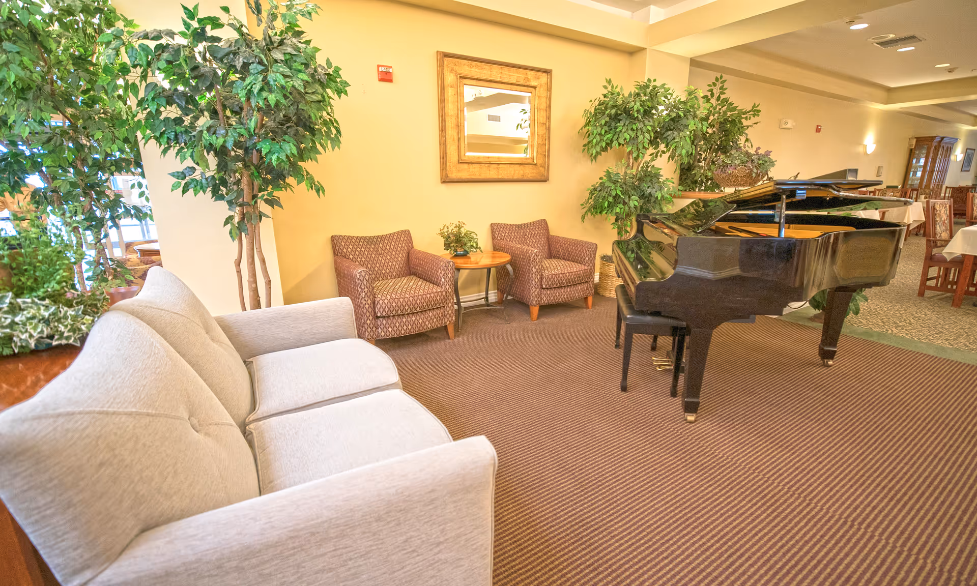 A spacious senior living lounge with a grand piano, beige seating, and potted trees.