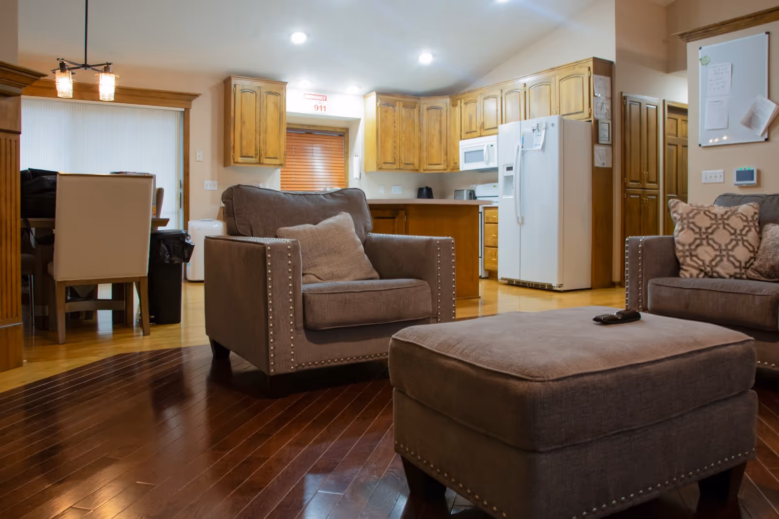 Open living room area with brown sofas, ottoman, and a nearby kitchen in warm wood tones.