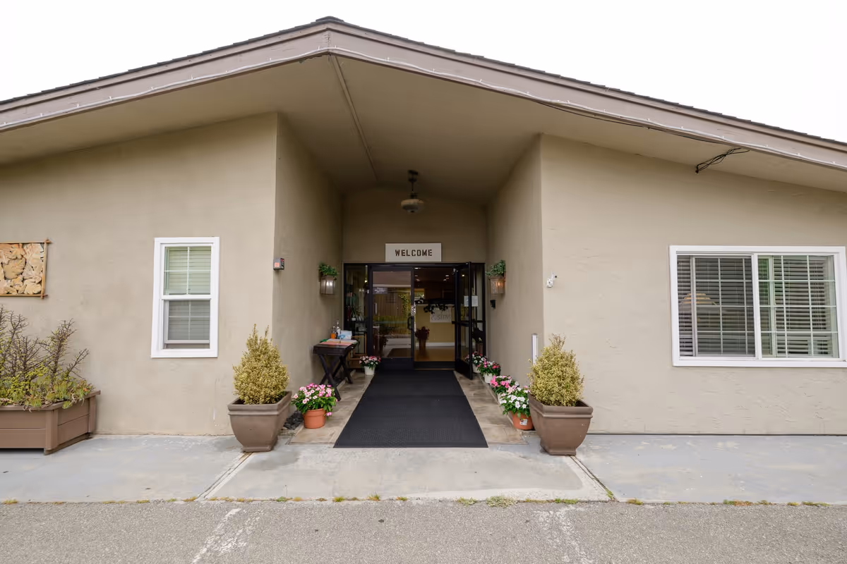 Beige Maple House senior living facility front entrance with a black mat and potted plants.