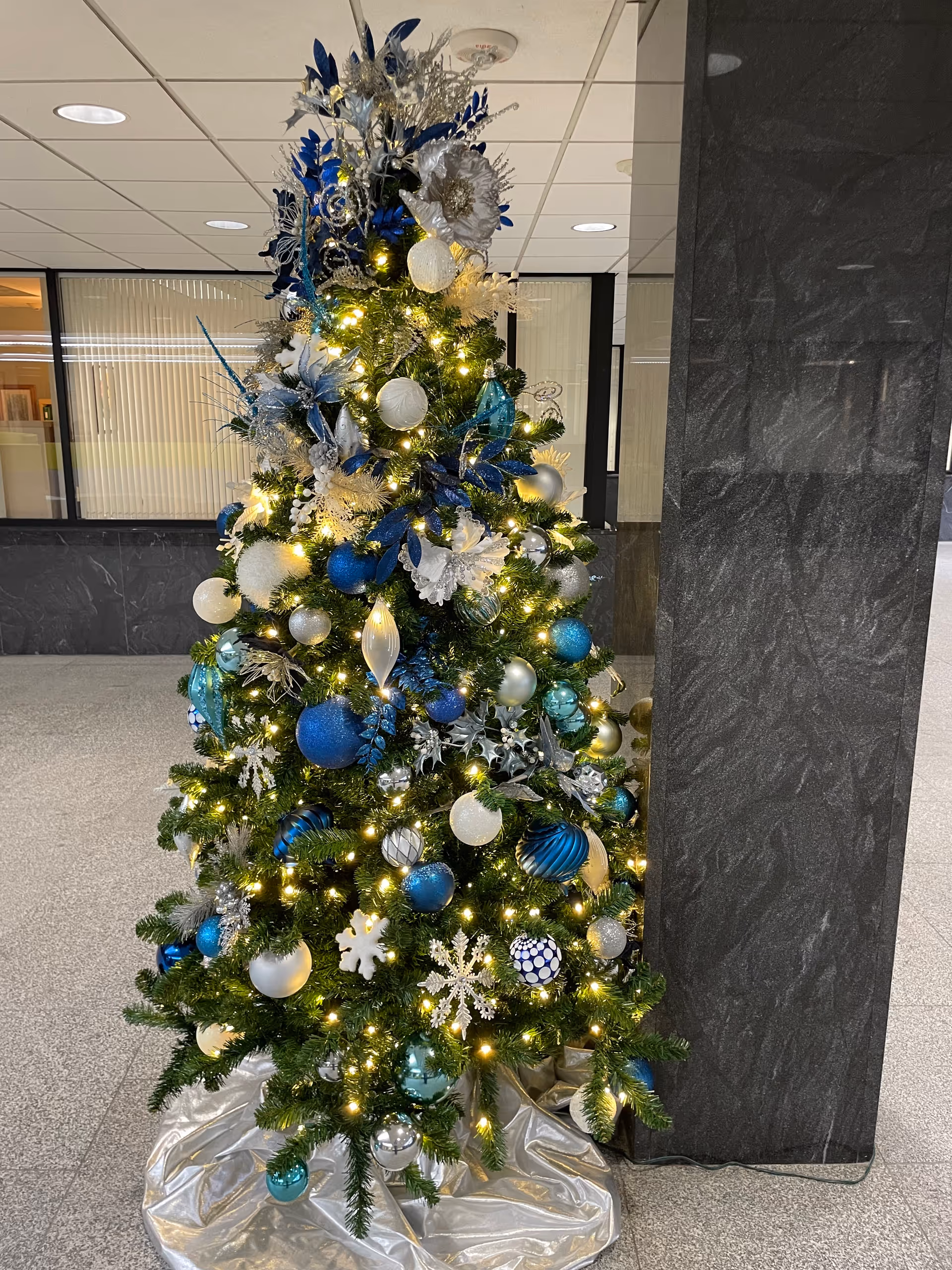 Decorated Christmas tree with blue, white, and silver ornaments indoors near a marble pillar.