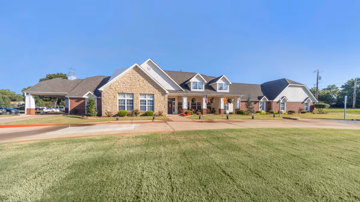 Exterior front view of a single-story senior living facility building with a combination of stone and brick facade, a covered entrance, and a well-maintained lawn under a clear blue sky.
