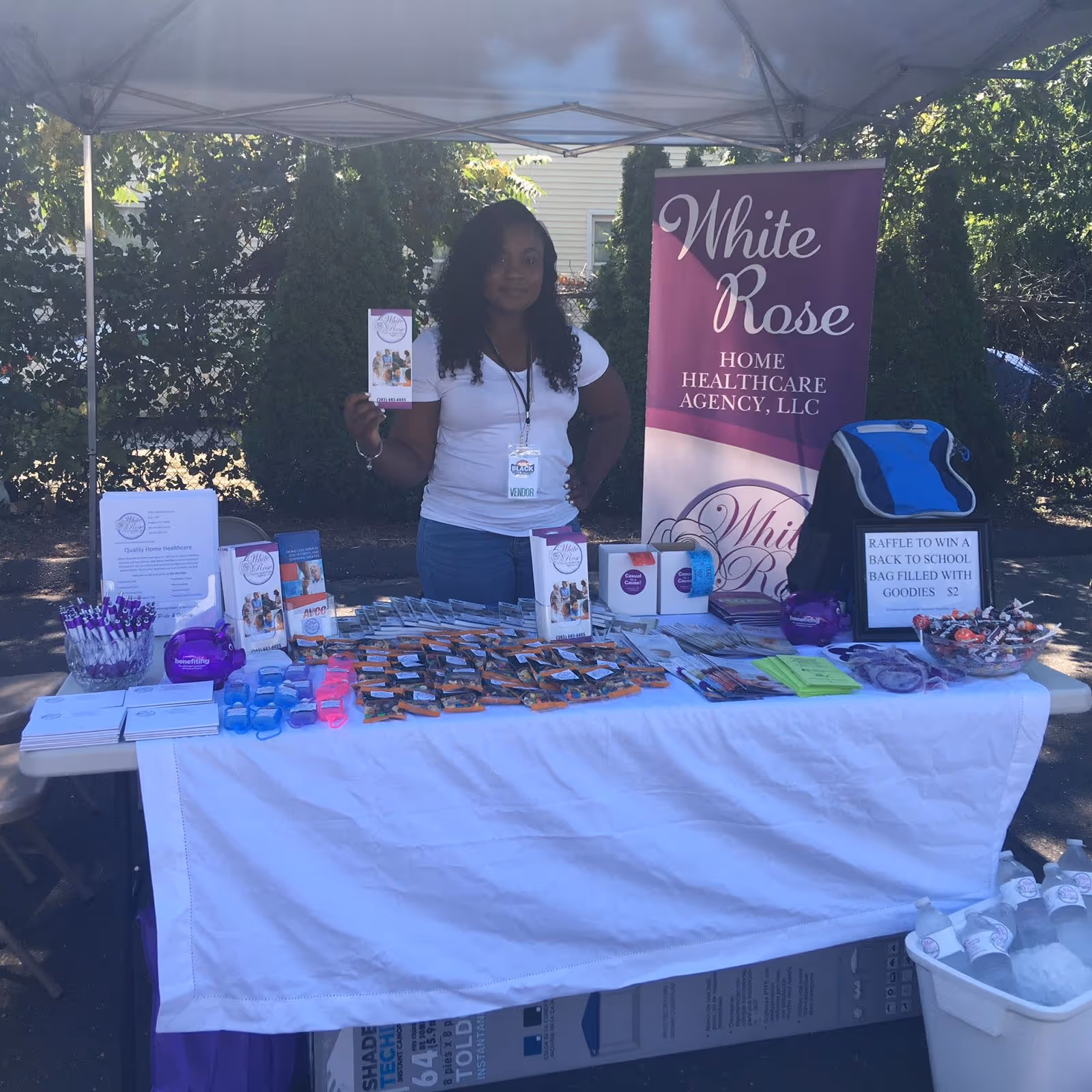 A woman stands behind a promotional table under a canopy at an outdoor event.