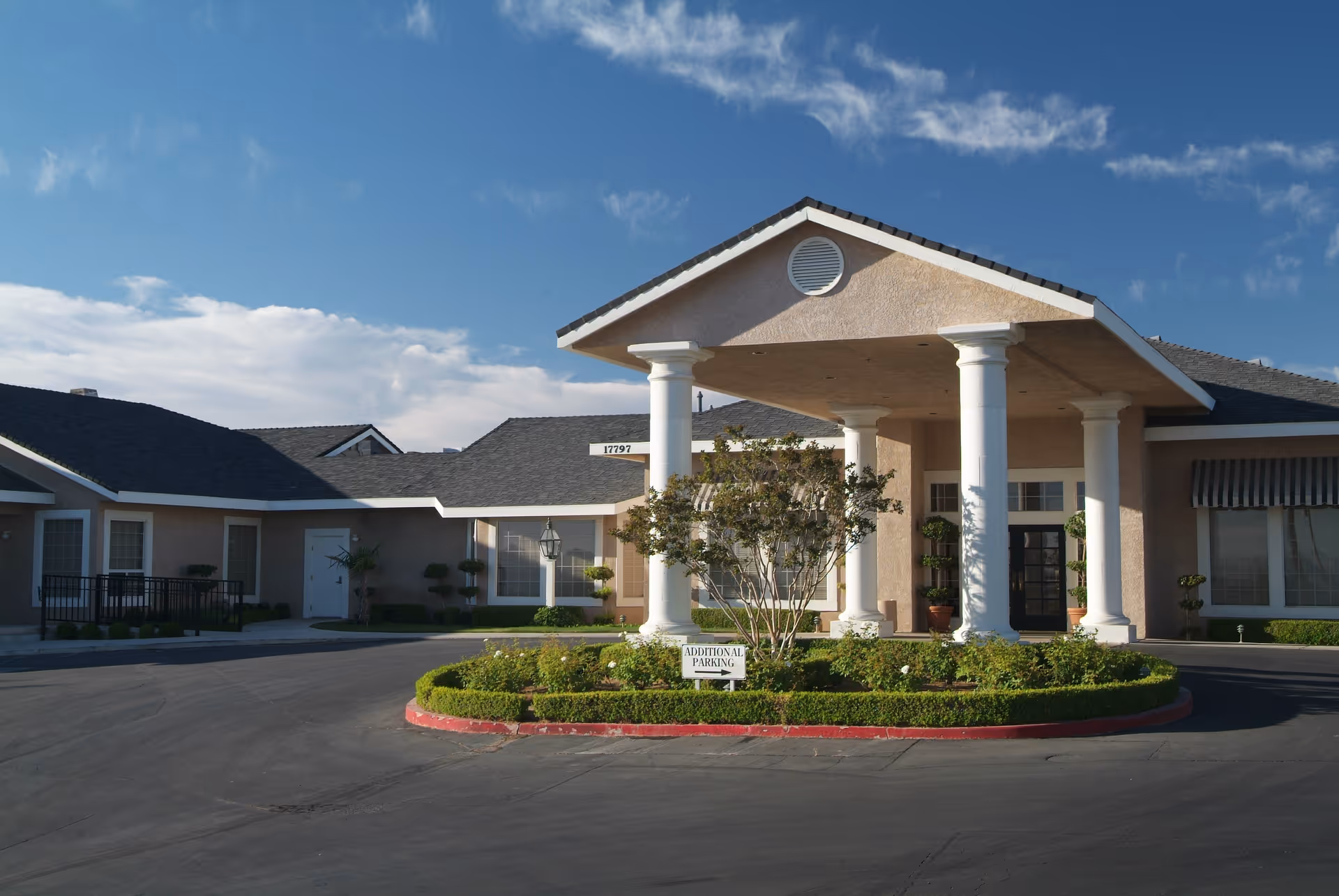 Front exterior view of Sterling Inn Assisted Living Community featuring a covered entrance with four white columns, manicured bushes, and a sign indicating additional parking. The building has beige walls, multiple windows, and a dark shingled roof under a blue sky with some clouds.