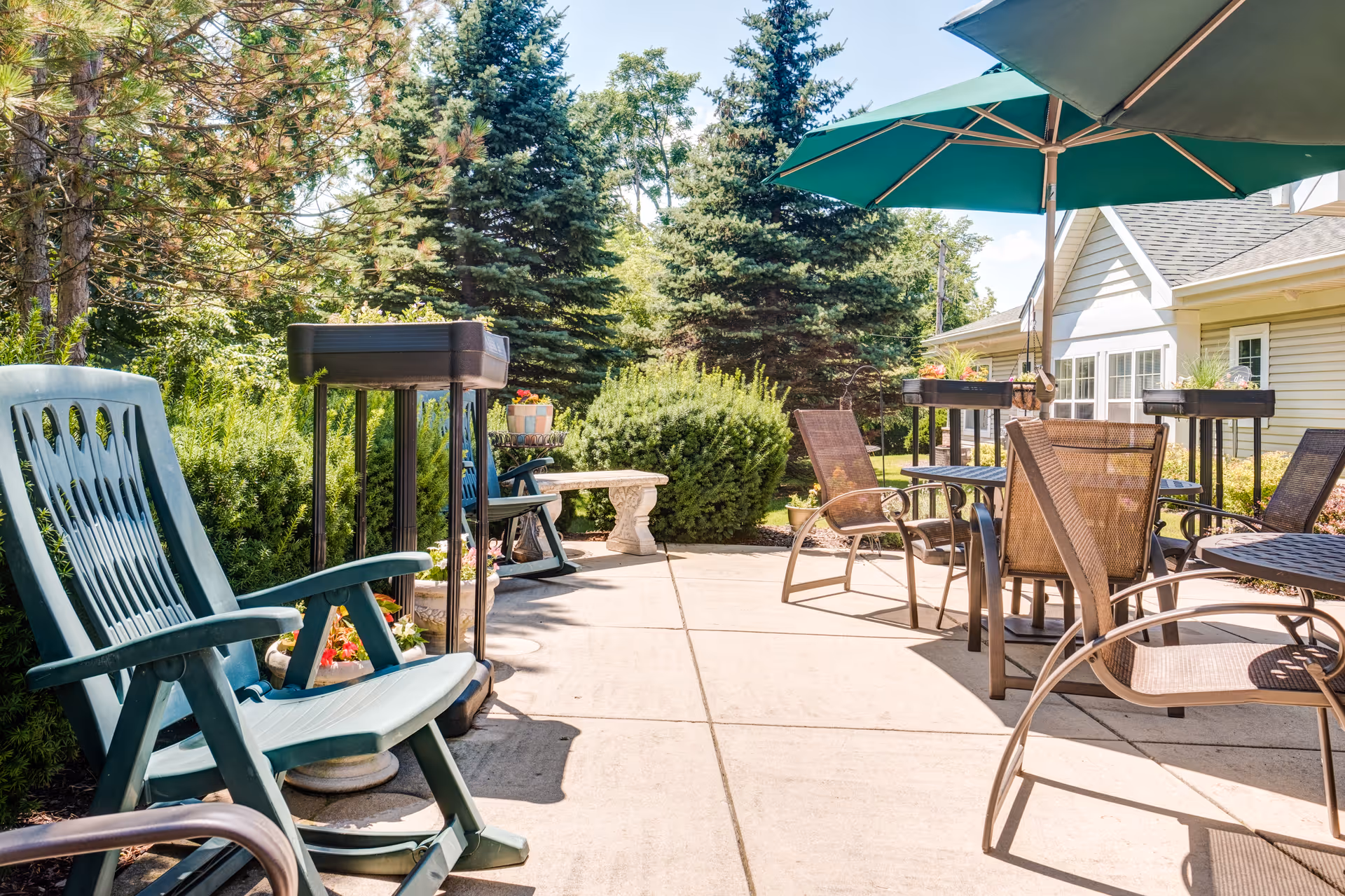 Outdoor patio area with several chairs and tables, including green plastic and brown mesh chairs, shaded by a large green umbrella. The patio is surrounded by lush greenery, bushes, and tall evergreen trees, with a light-colored building visible in the background under a clear blue sky.