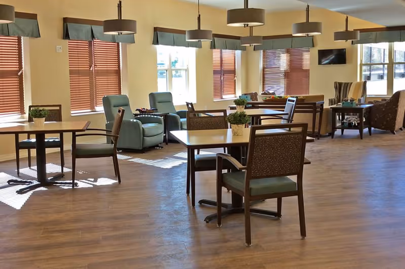 A spacious, well-lit common area with multiple tables and chairs arranged for seating. There are several armchairs and a sofa near the windows with wooden blinds. The room has wooden flooring, beige walls, and modern ceiling lights. Small potted plants are placed on the tables.