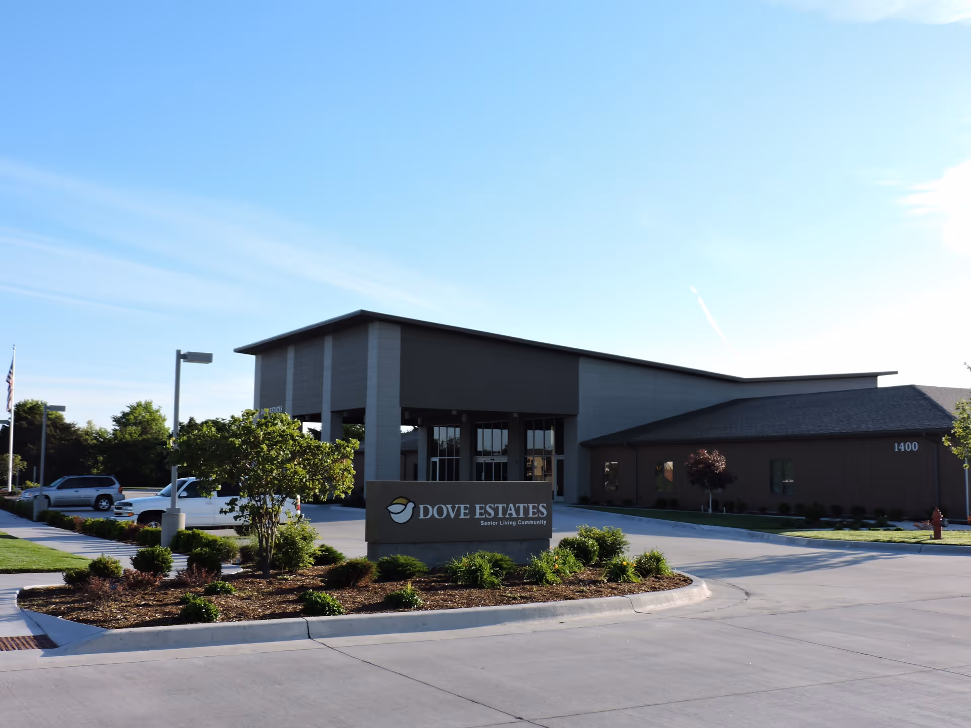 Exterior view of Dove Estates Senior Living Community building with a clear blue sky, landscaped greenery, and a parking area with several vehicles.