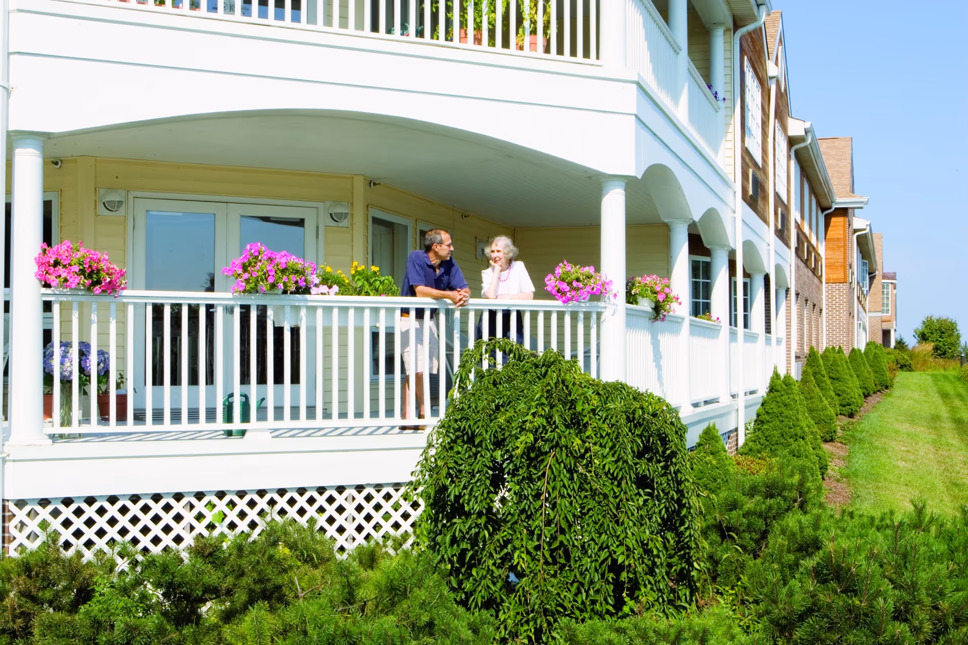 Two elderly people talking on a flower-lined balcony of a multiunit residential building.