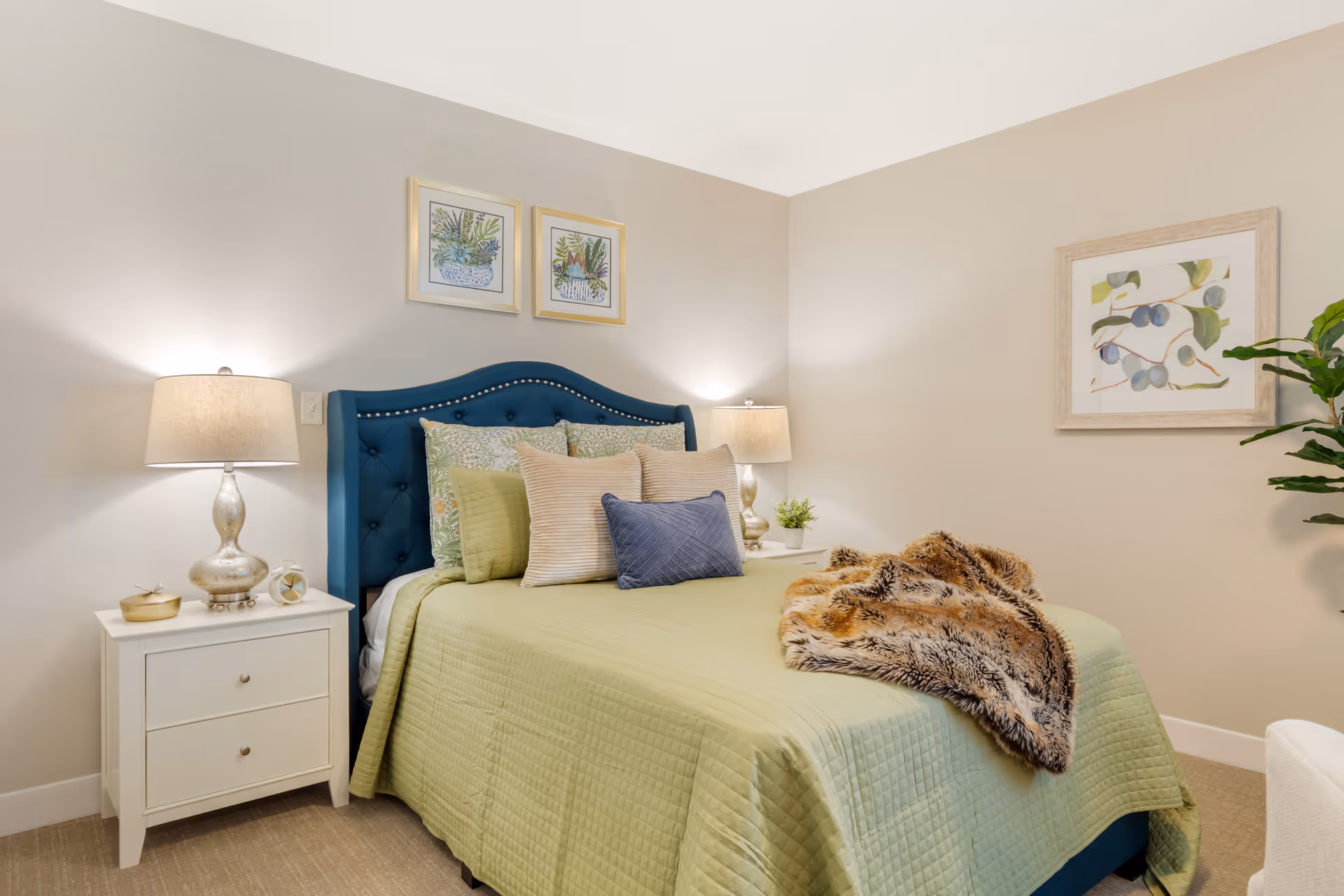 Bright bedroom with green quilt, blue upholstered headboard, twin nightstands, lamps, and botanical wall art.