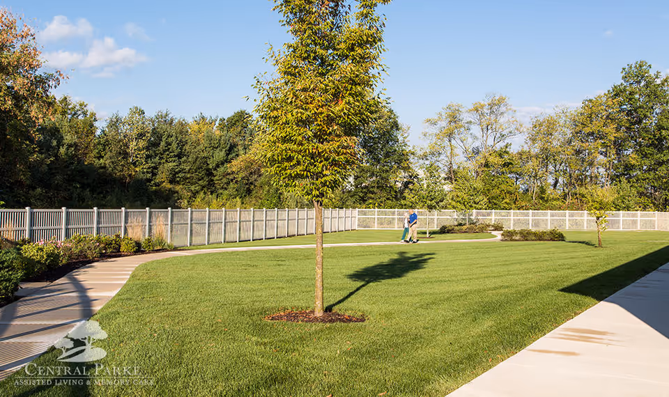 A well-maintained outdoor garden area with a curved concrete walkway, green grass, a few small trees, and a white fence enclosing the space. Two people are walking along the path in the distance under a clear blue sky.