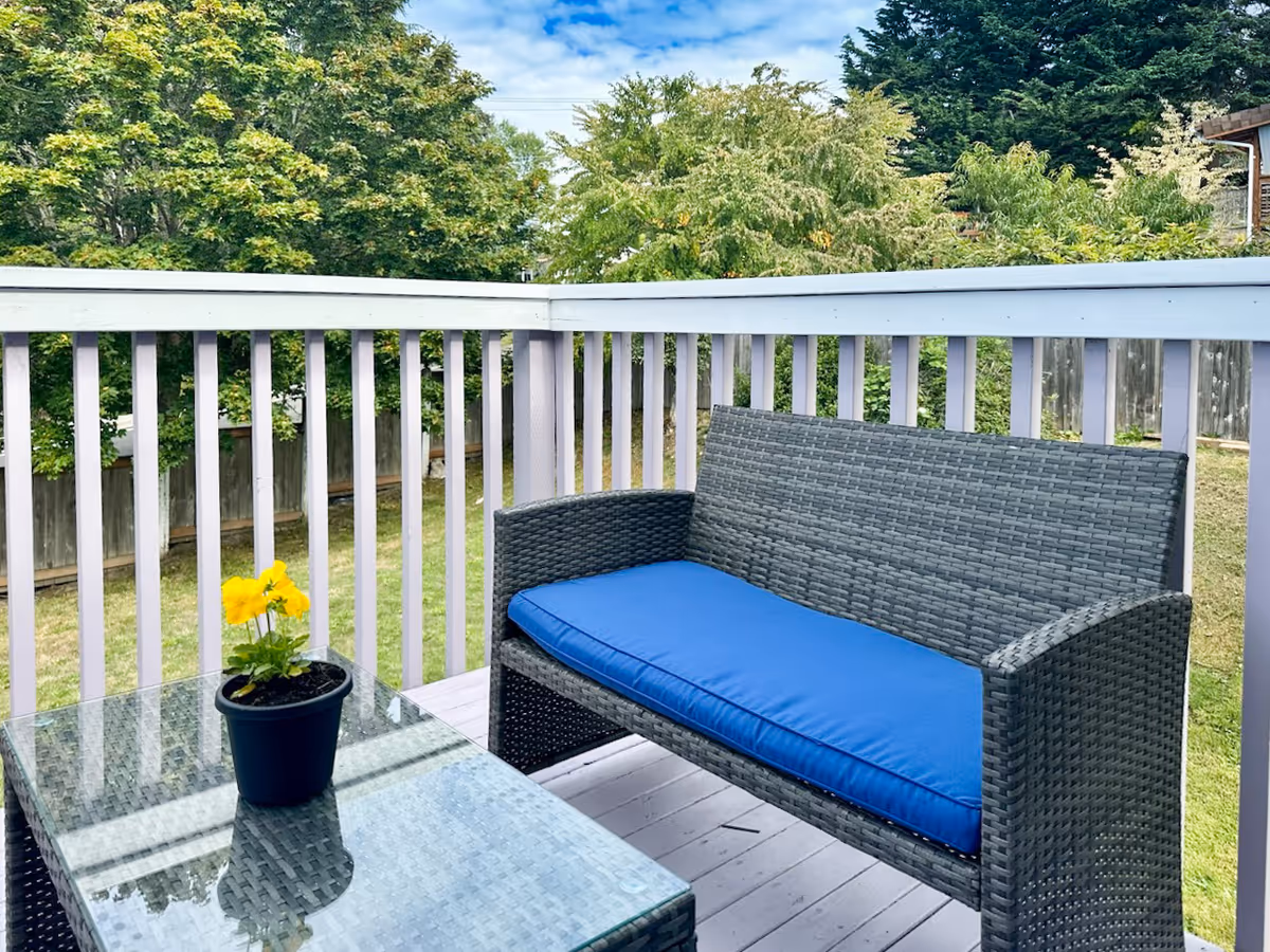 Outdoor balcony with wicker sofa, glass table, and a yellow potted flower.