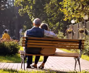 An elderly couple sits on a wooden bench beside a sunlit park path outdoors.