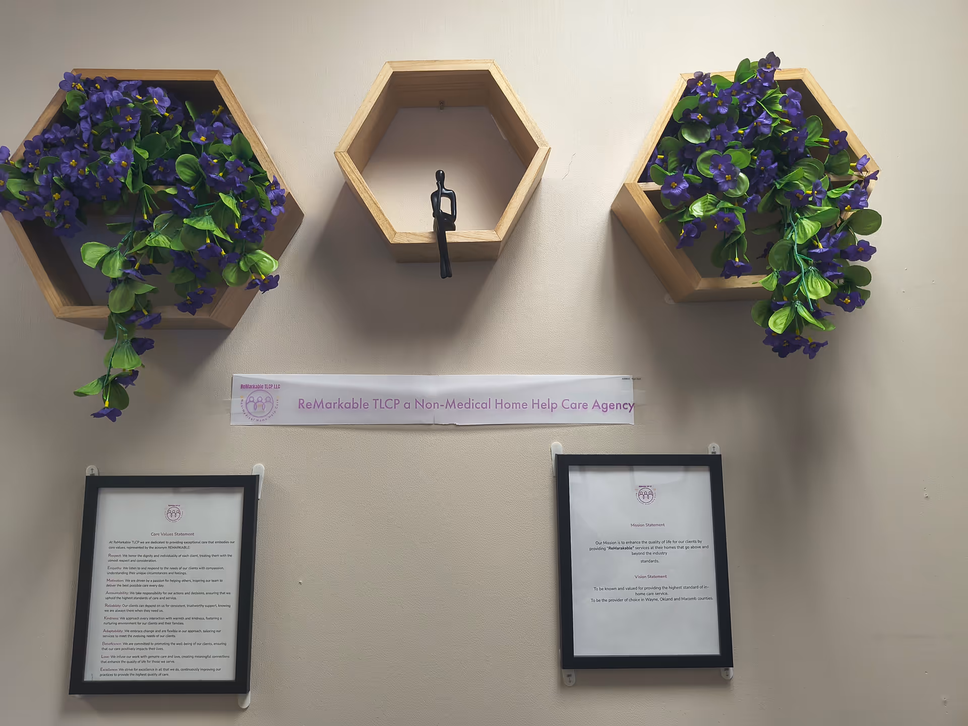 Interior wall with hexagonal planters, purple flowers, and framed documents beneath a banner.