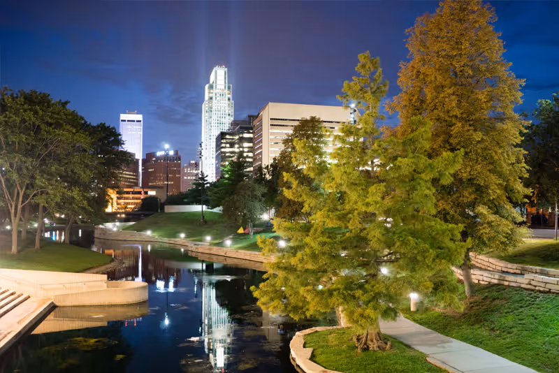 Nighttime exterior view of a river park with trees and city buildings.