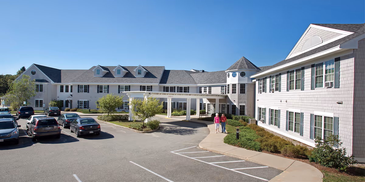 Front exterior of a large white two-story senior living building with a covered entrance, parking lot, and two people walking on the sidewalk.