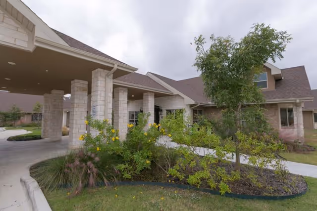 Front entrance of a senior living facility with a covered porte-cochere, stone columns, and landscaped shrubs.