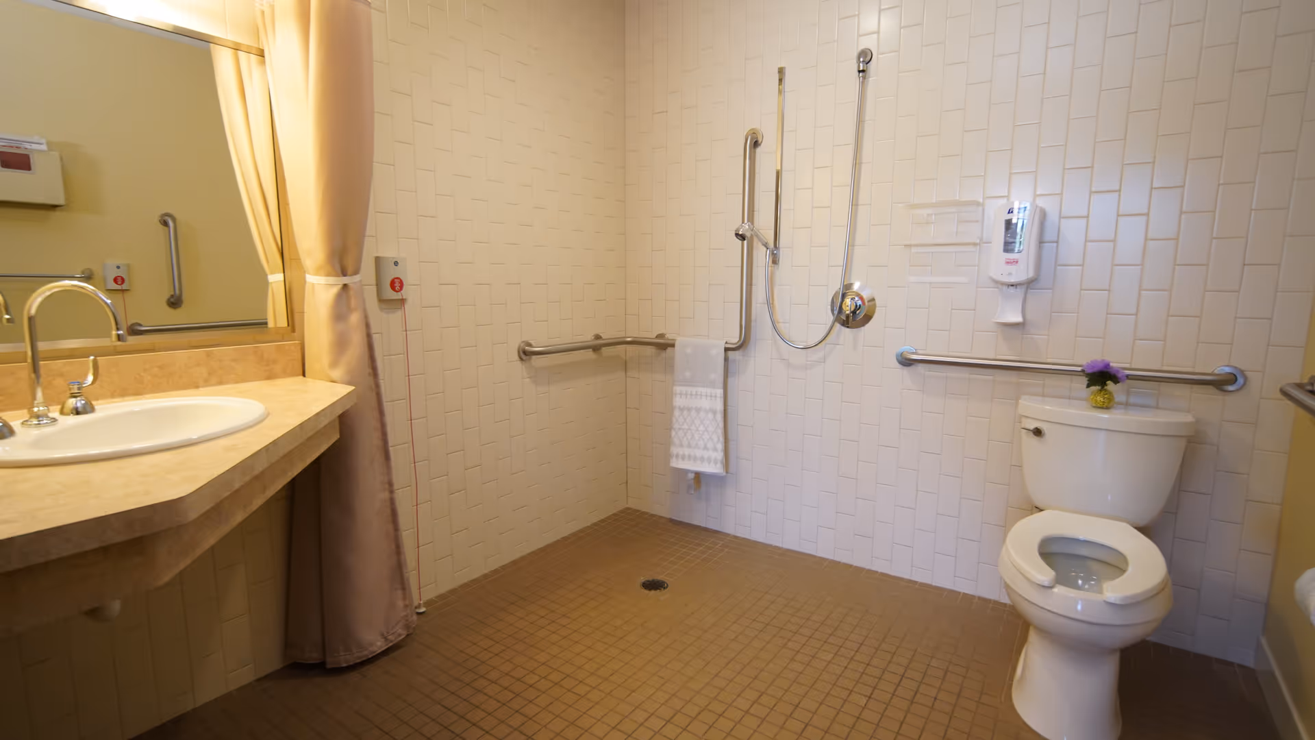 Accessible bathroom with beige tiled floor and white tiled walls. The bathroom features a toilet with a small purple flower on top of the tank, a wall-mounted soap dispenser, grab bars around the toilet and shower area, a handheld showerhead, a towel hanging on a grab bar, a sink with a countertop, and a large mirror partially covered by a beige privacy curtain.