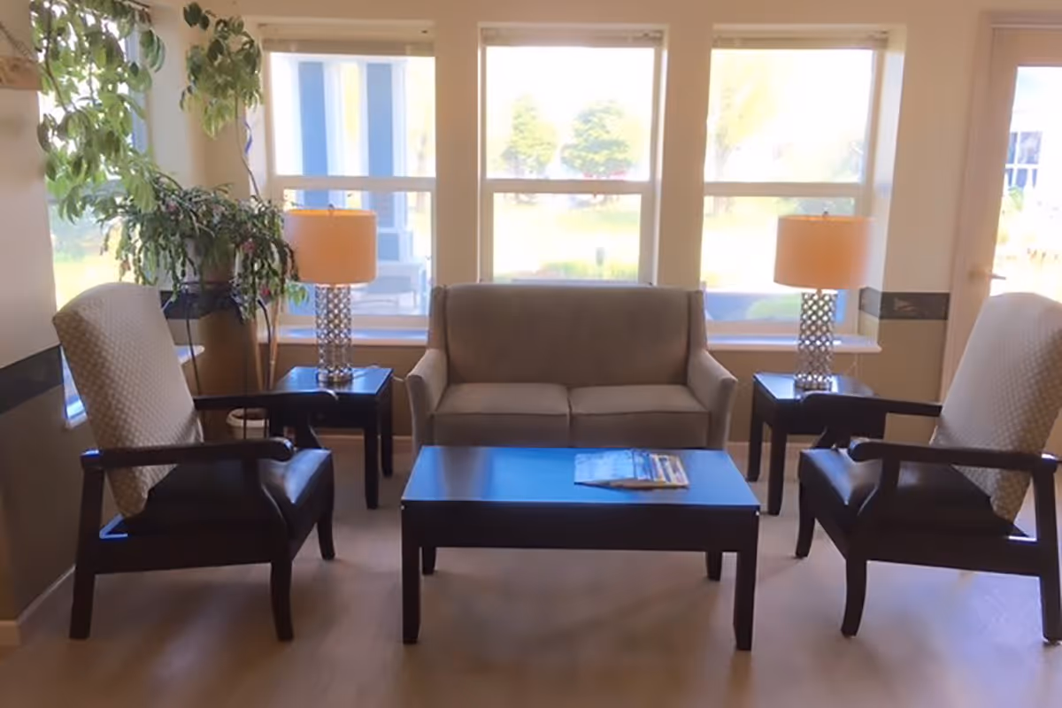 A cozy living room area with a gray sofa centered between two armchairs with patterned cushions. There are two side tables with matching lamps on either side of the sofa. A coffee table with magazines is in front of the sofa. Large windows behind the sofa let in natural light and show a view of greenery outside. A tall potted plant is placed near the left side of the sofa.