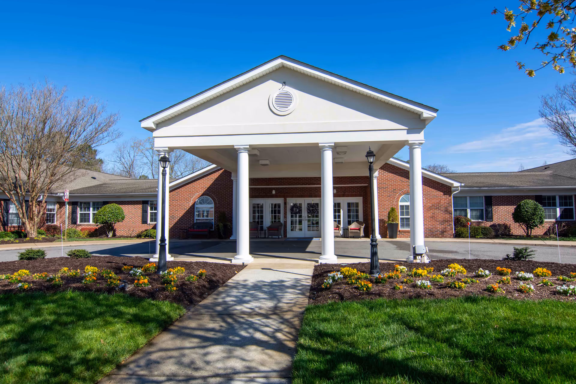 Brick senior living facility entrance with a portico, white columns, and landscaped walkway.