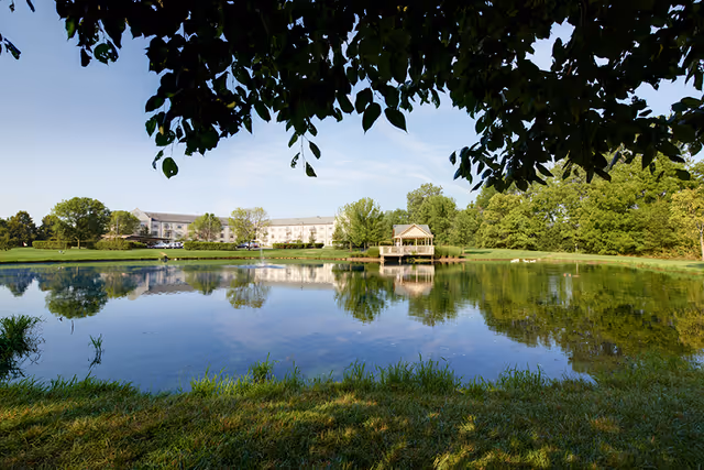 A serene outdoor scene featuring a large pond with clear reflections of trees and a gazebo on the water. In the background, there is a multi-story building surrounded by greenery under a clear blue sky. The image is framed by tree branches and leaves at the top.