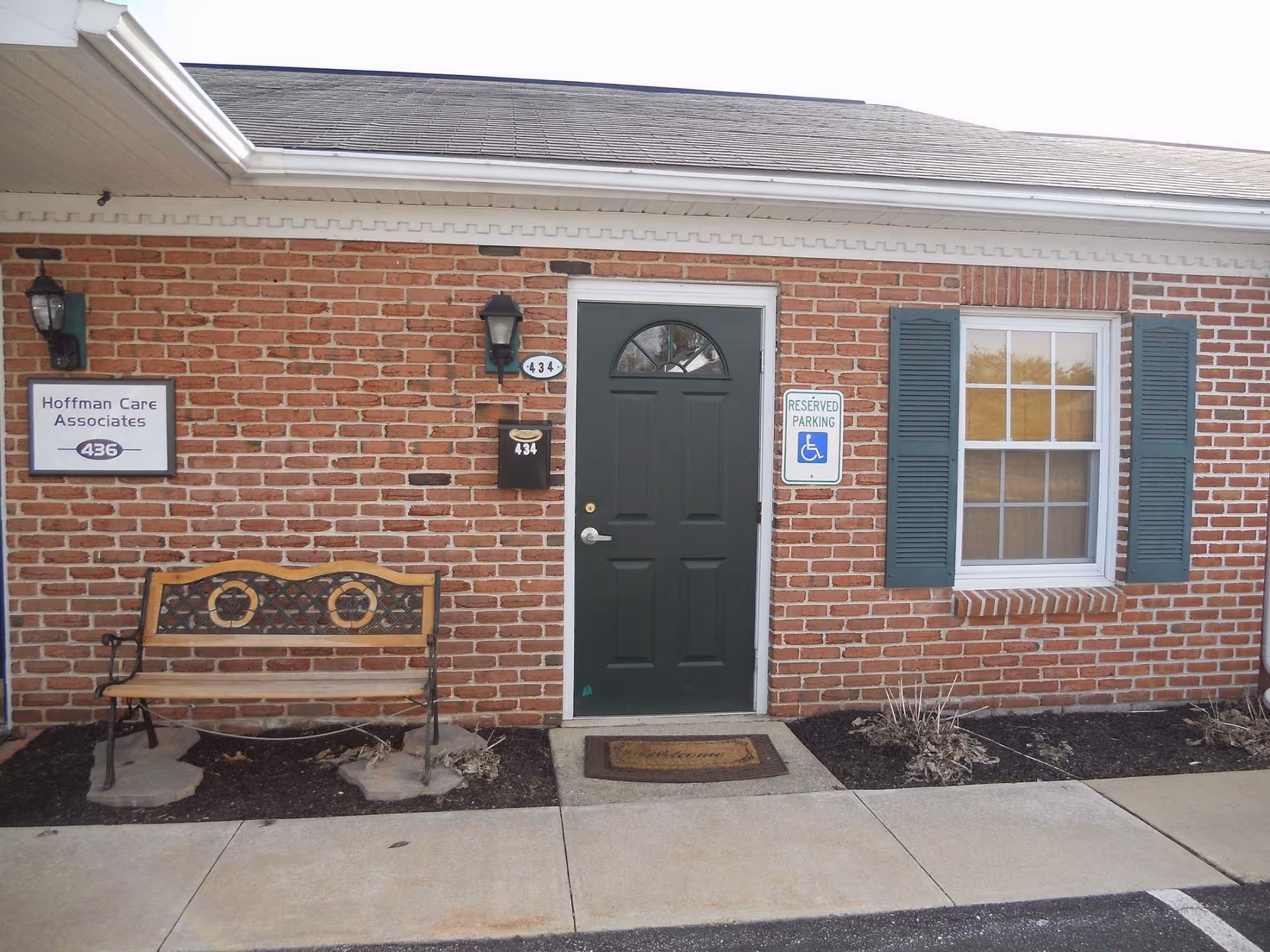 Brick exterior of Hoffman Care Associates facility with green door, bench, and handicap parking sign.