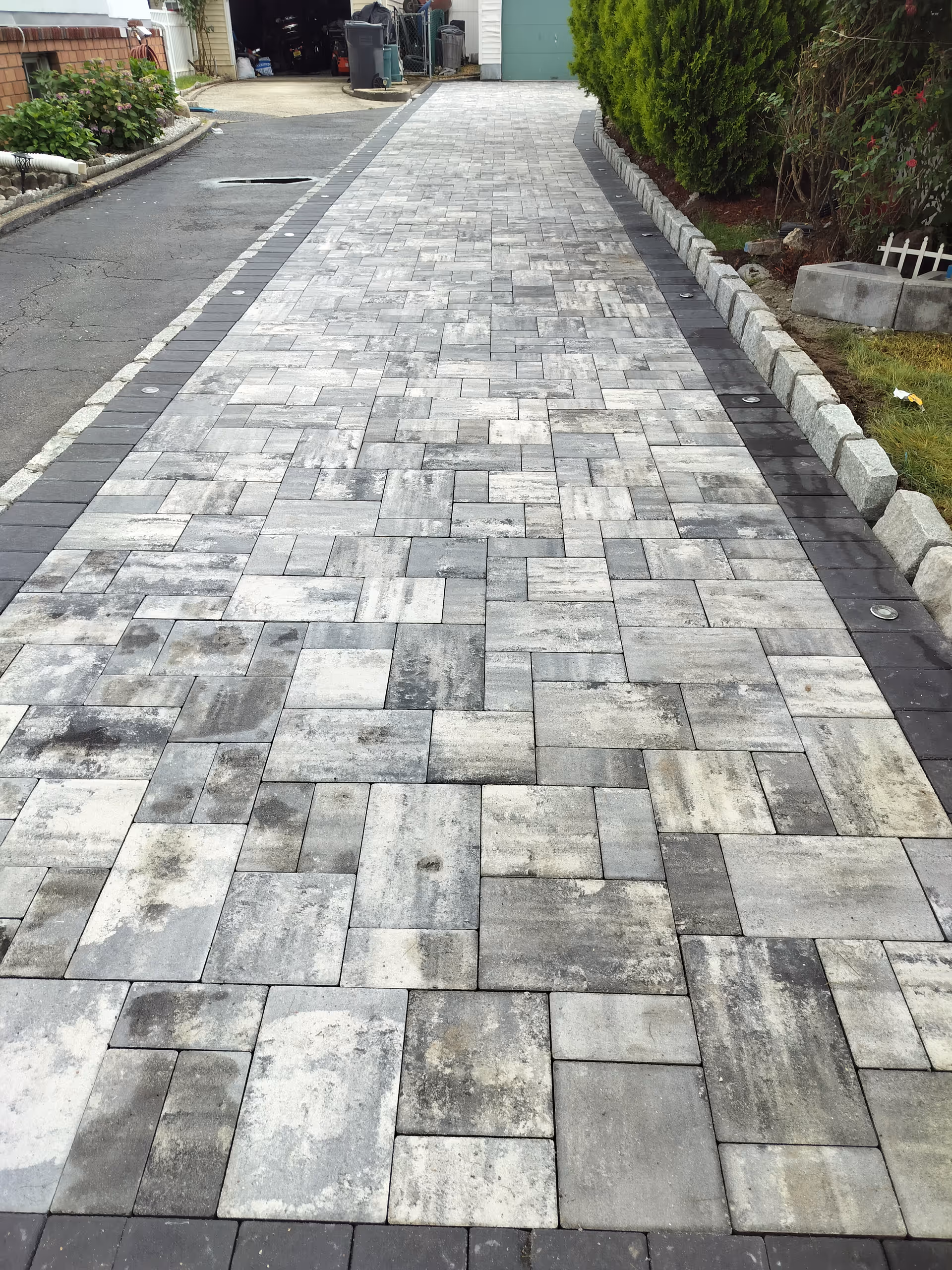 Wide outdoor brick-paver walkway toward a garage with surrounding greenery.