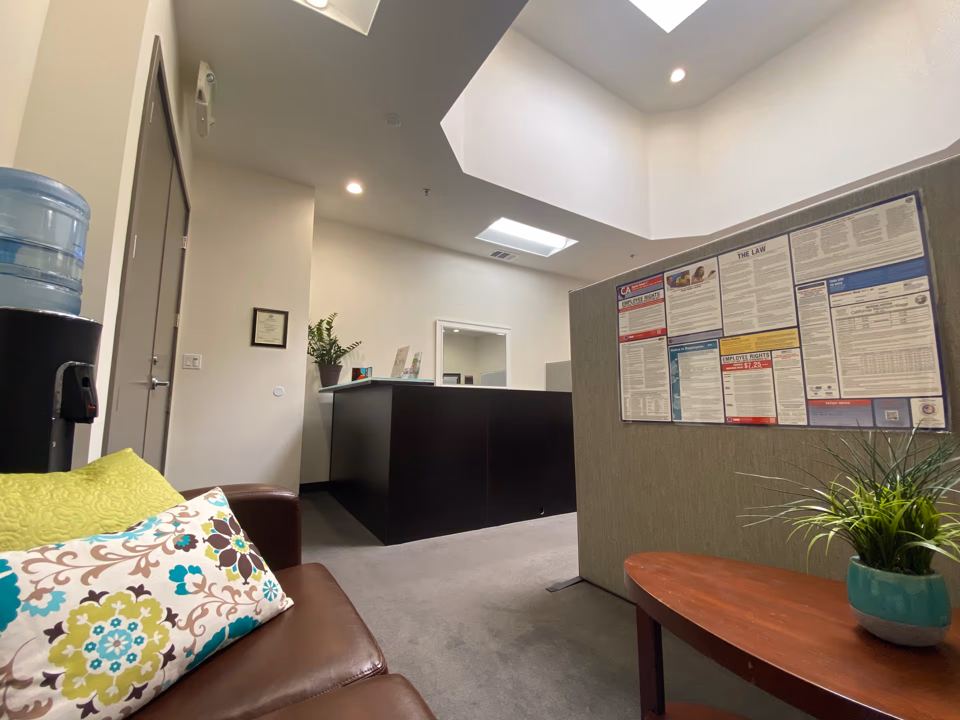 Reception lobby with dark desk, a couch with patterned pillows, bulletin board, water cooler, plants.
