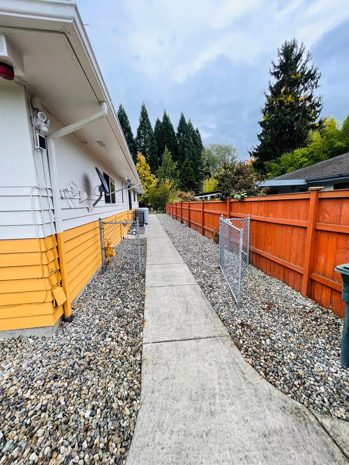Narrow concrete walkway beside a yellow and white facility, with a red wooden fence and gravel.
