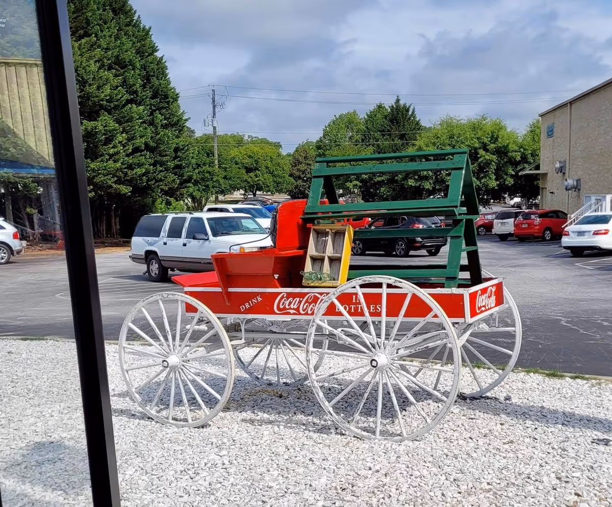 A vintage Coca-Cola cart sits on gravel in a parking lot near a building.