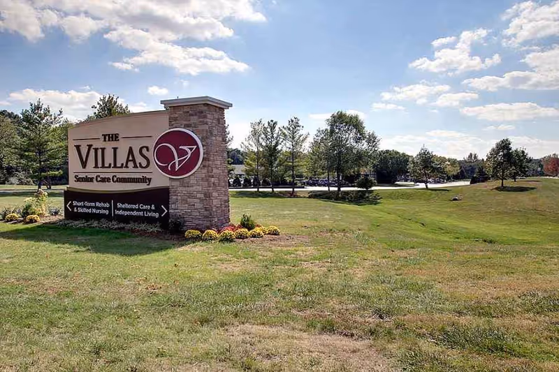 Stone entrance sign reading 'The Villas Senior Care Community' on a grassy lawn with trees and a blue sky.