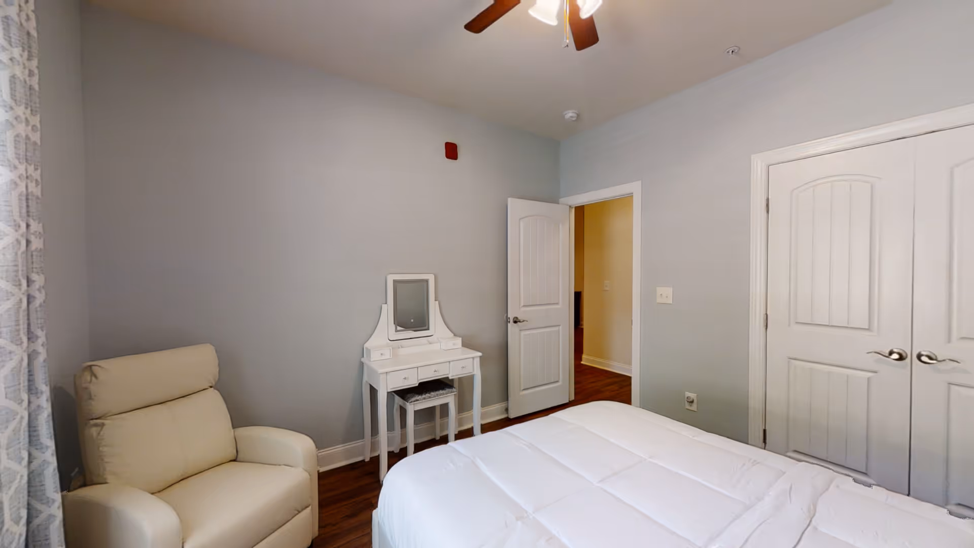 A bedroom in a senior living facility features a bed, armchair, and vanity.