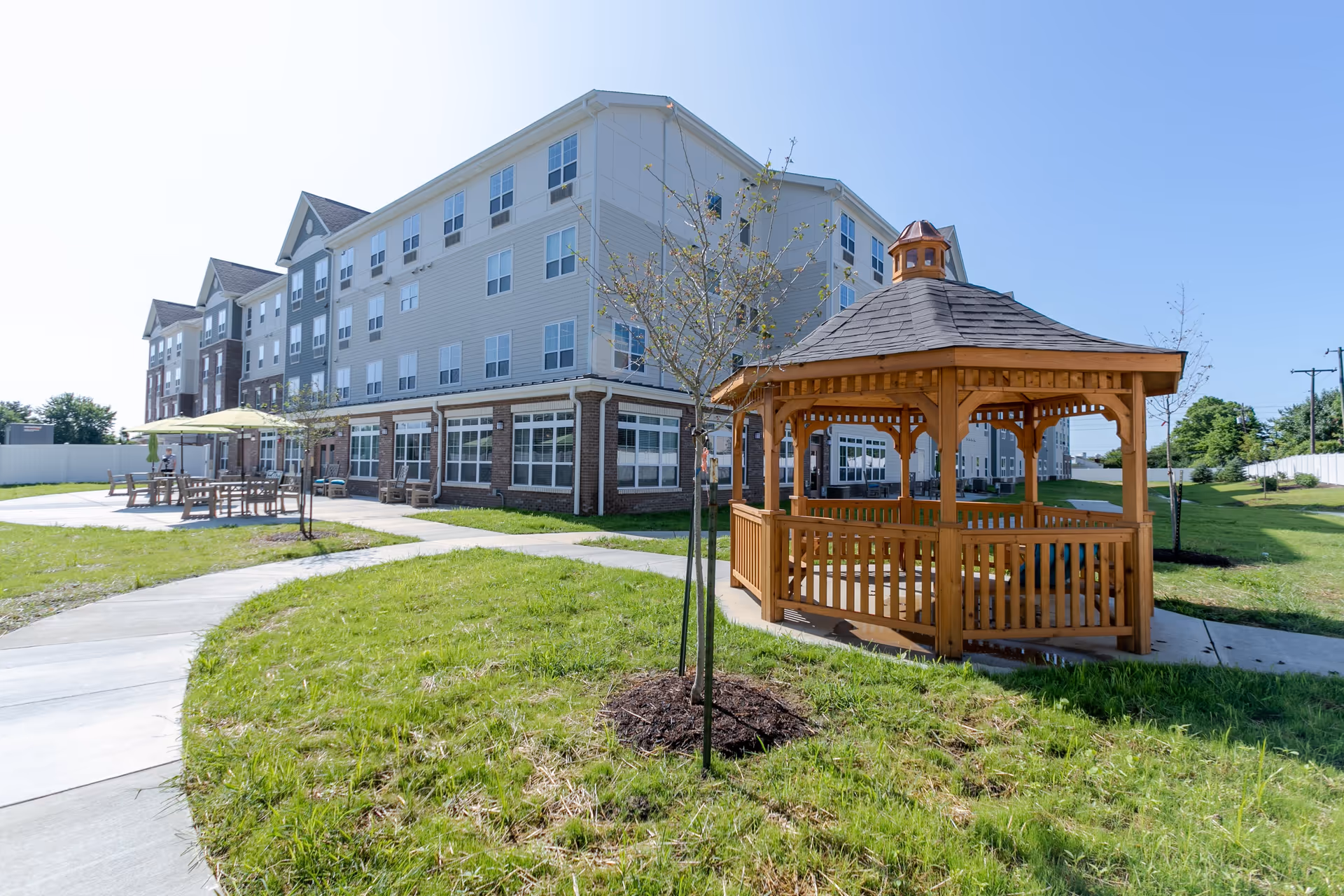 Outdoor area of a senior living facility featuring a wooden gazebo, a paved walkway, young trees, and patio tables with umbrellas. The multi-story building with numerous windows is visible in the background under a clear blue sky.