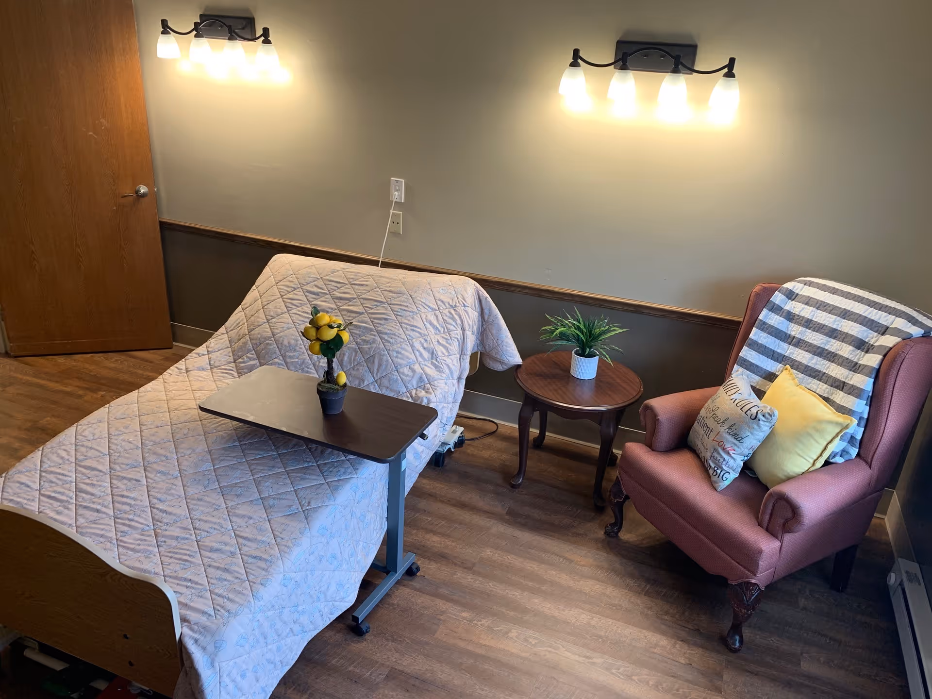 A cozy room with a hospital-style bed covered with a quilted blanket, a small overbed table with a decorative lemon tree, a round wooden side table with a potted plant, and a comfortable upholstered armchair with two pillows and a striped throw blanket. The room has wooden flooring, beige walls with a wooden trim, two wall-mounted light fixtures above the bed, and a closed wooden door.
