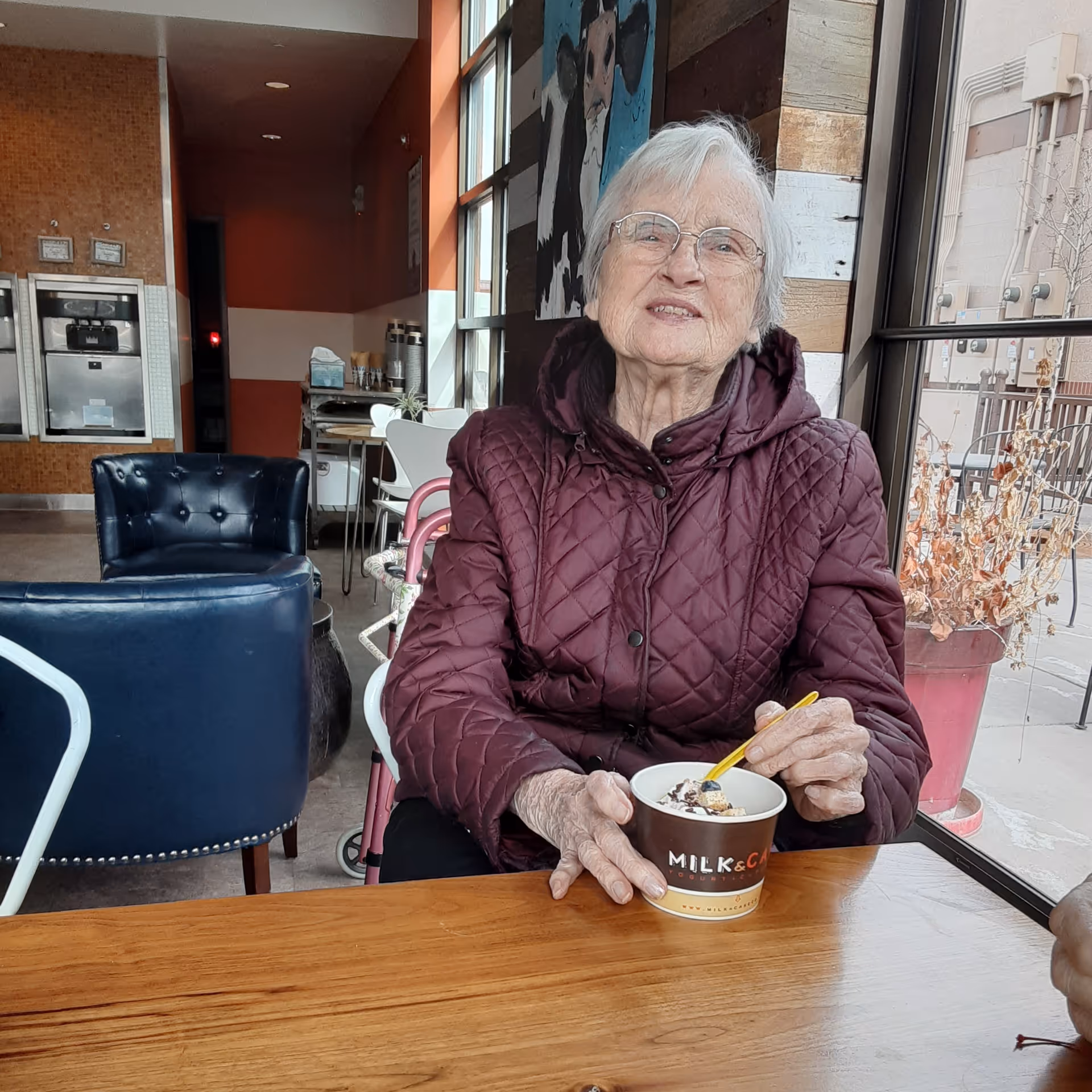 An elderly woman wearing glasses and a maroon quilted jacket is sitting at a wooden table inside a cafe or dining area. She is holding a cup of ice cream or dessert with a yellow spoon. Behind her, there are blue chairs, a window showing an outdoor patio with dried plants, and a wall with a painting of a dog.