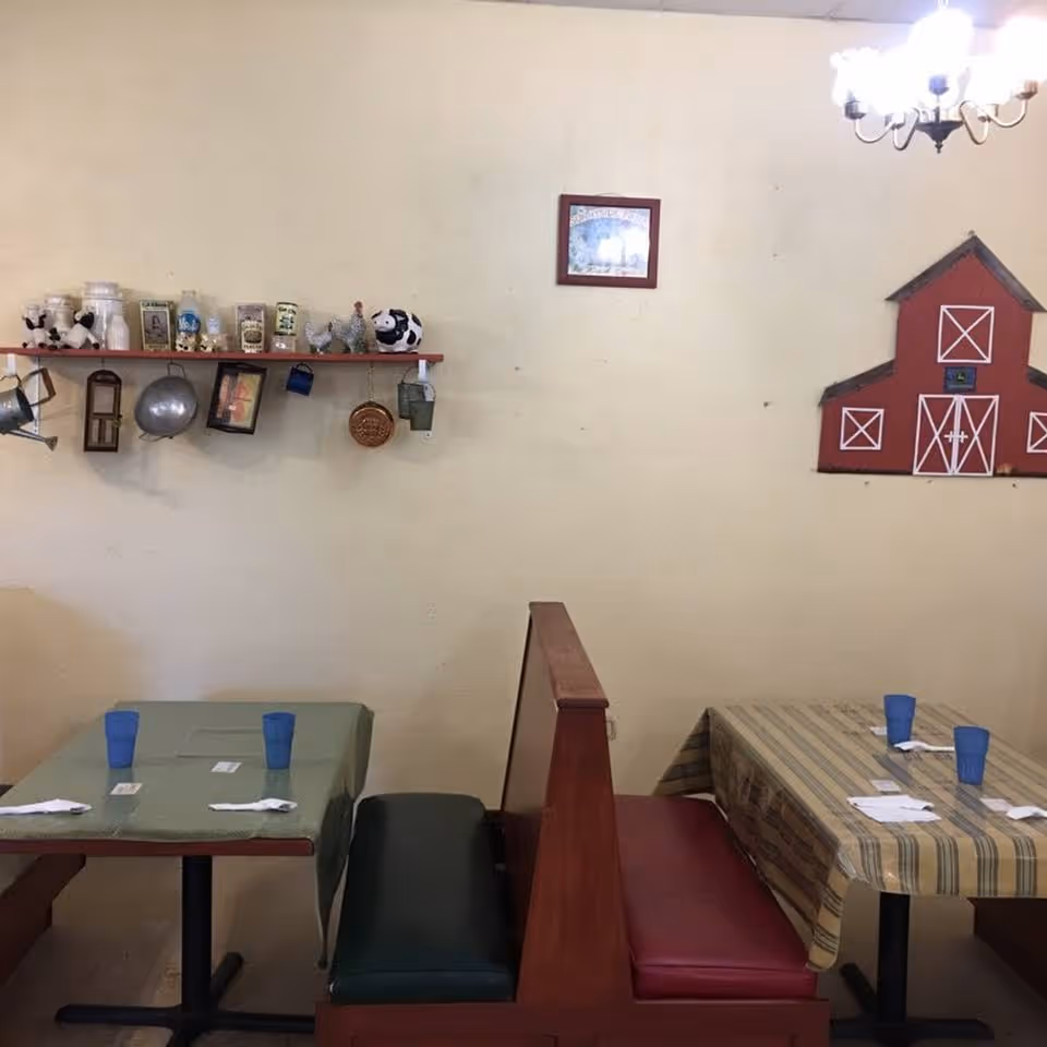 Interior view of a dining area with two tables covered with tablecloths, each set with blue plastic cups and napkins with utensils. The seating consists of booth benches with green and red cushions. On the wall above, there is a shelf holding decorative items including jars, a watering can, and animal figurines. A red barn-shaped wall decoration and a framed picture are also visible. A chandelier hangs from the ceiling.