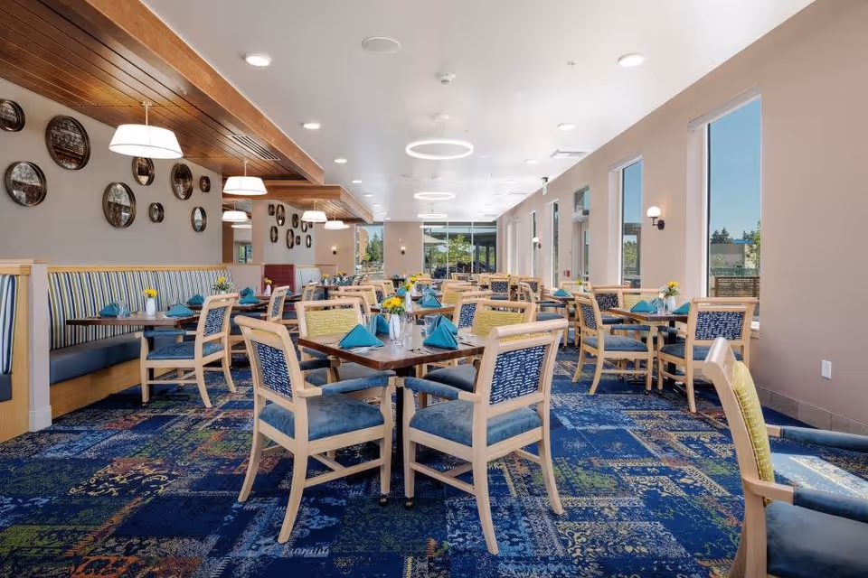 A bright and spacious dining room with multiple wooden tables and chairs arranged neatly. Each table is set with teal napkins and small flower vases. The room features large windows on one side allowing natural light to fill the space, modern circular ceiling lights, and decorative round mirrors on the wall. The carpet has a blue patterned design, and the overall ambiance is clean and welcoming.