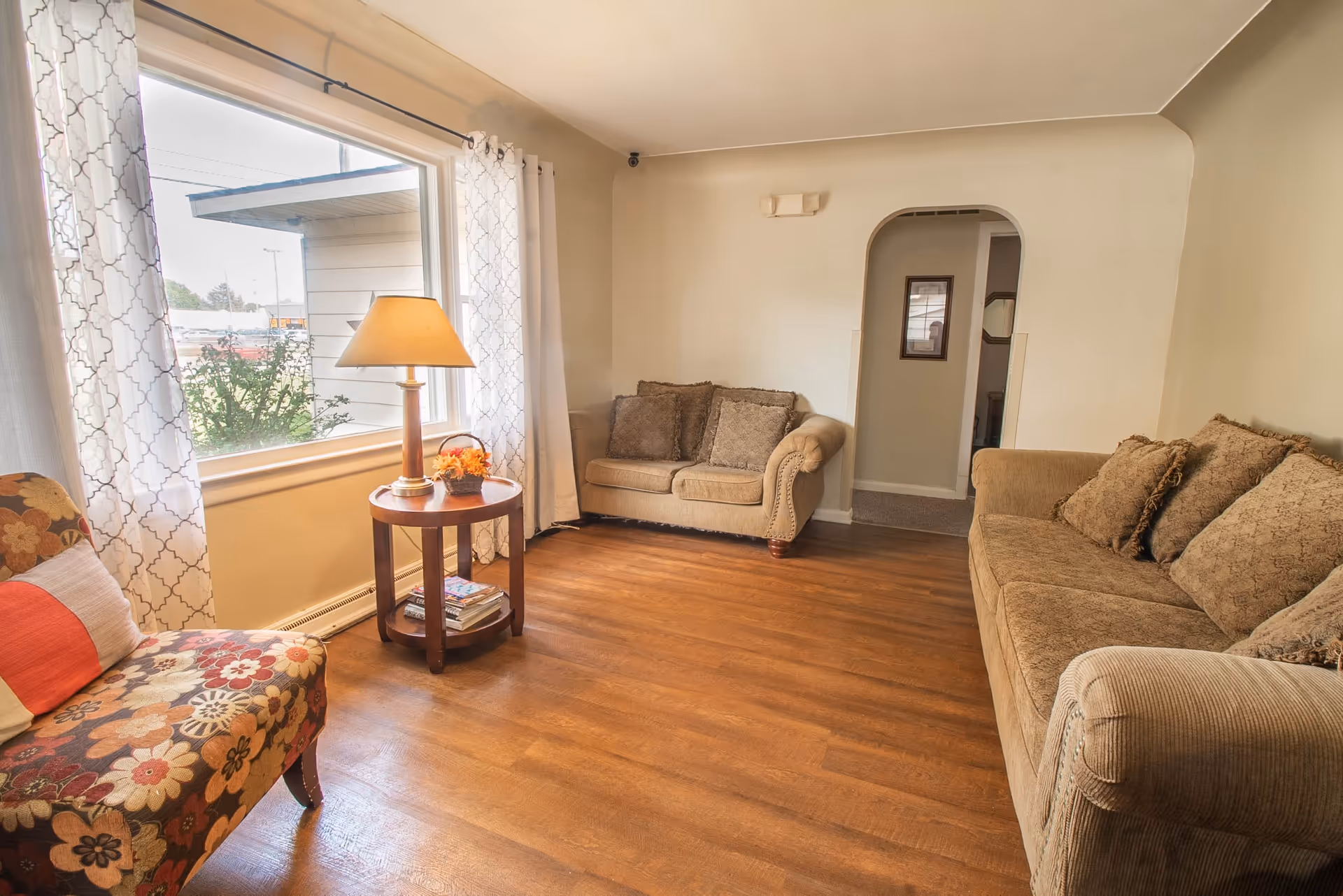 A cozy living room with wooden flooring, two beige sofas with patterned cushions, a floral upholstered chair with a red and white pillow, a round wooden side table with a lamp and a small flower arrangement, and a large window with white patterned curtains letting in natural light.