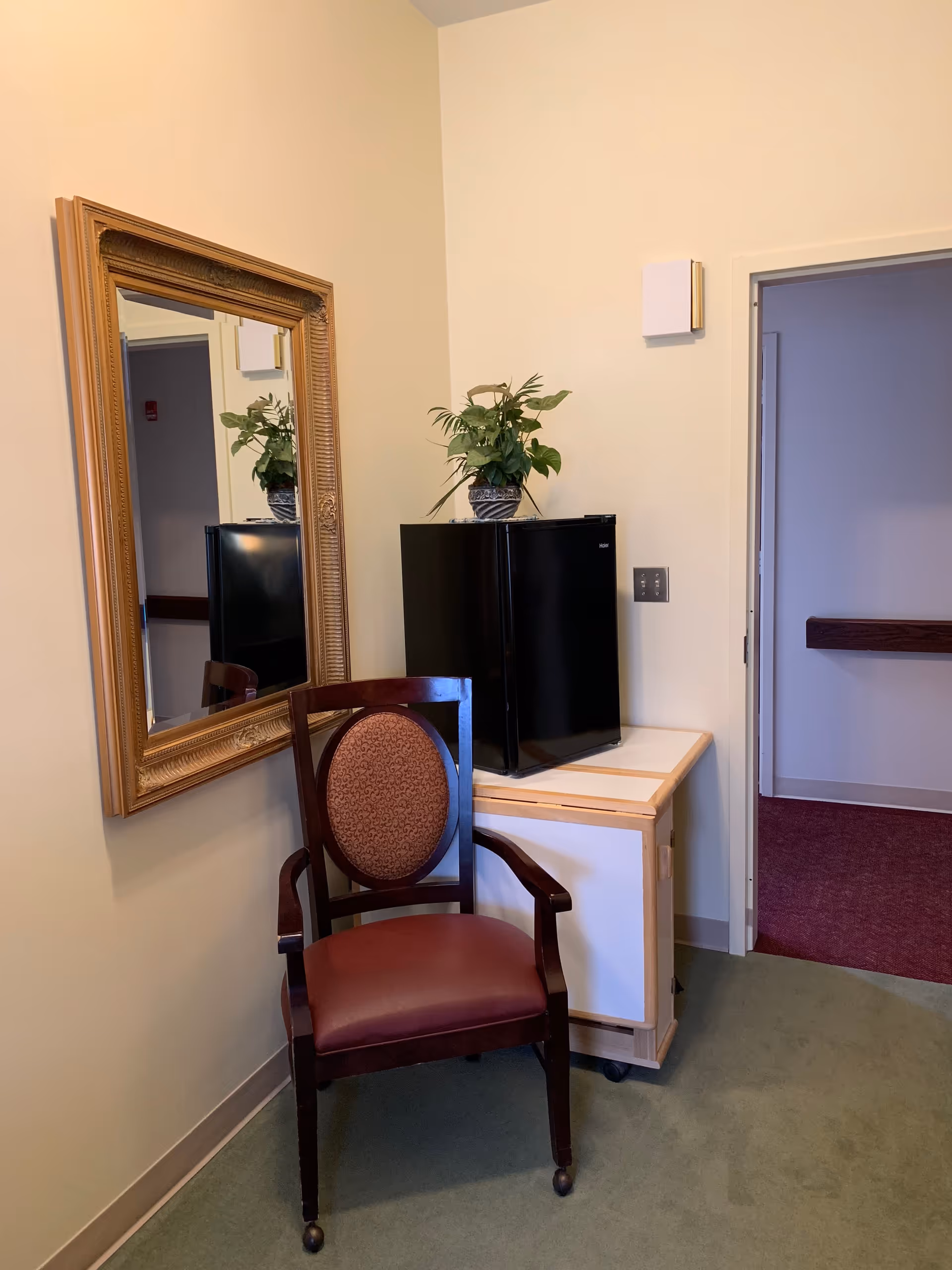 A corner of a room with a wooden chair featuring a cushioned seat and backrest, a small black mini fridge on a wooden cabinet with wheels, a potted plant on top of the fridge, and a large ornate gold-framed mirror hanging on the wall. The room has light-colored walls and green carpet, with an open doorway leading to a hallway with red carpet.