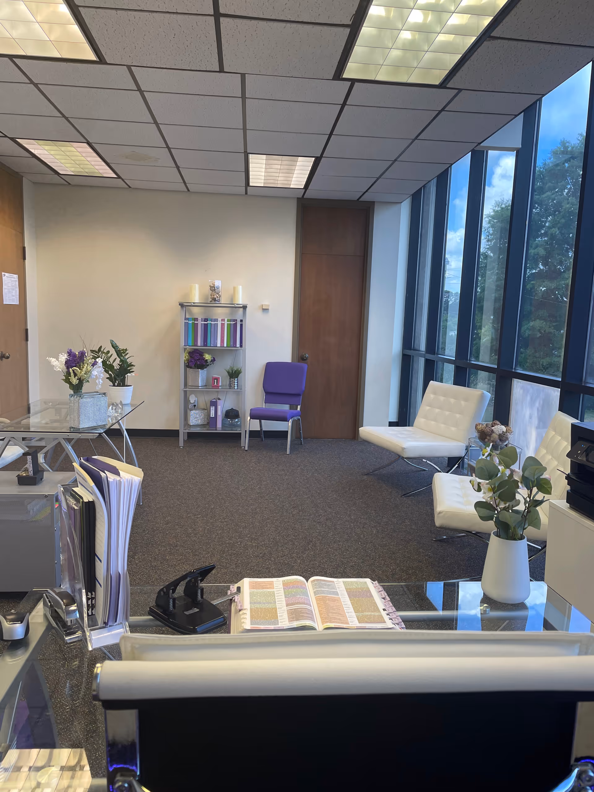 Interior reception area with glass desk, white seating, purple chairs, and large window wall.
