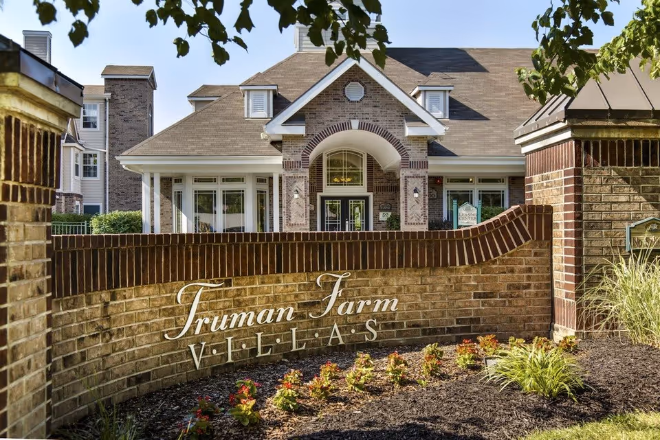 Entrance view of Truman Farm Villas showing a brick wall with the facility name in white letters, surrounded by landscaped plants and flowers, with the building's front entrance visible in the background under a clear sky.