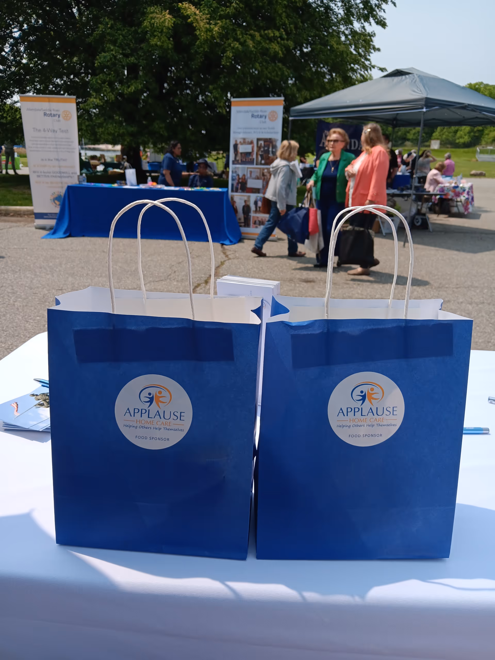 Two blue Applause Home Care bags sit on a table at an outdoor event.