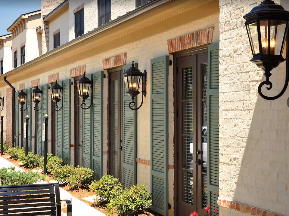 Row of green-shuttered French doors with wall lanterns and landscaping along a light-brick building exterior.