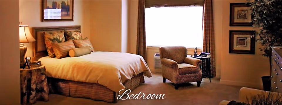A cozy bedroom with a neatly made bed featuring floral and striped pillows, a bedside table with a lamp, an armchair near a window with curtains, framed artwork on the walls, and a dresser with a plant beside it.