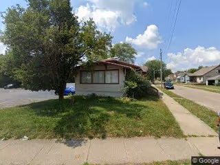 A small single-story building with a lawn, tree, and sidewalk along a quiet street.