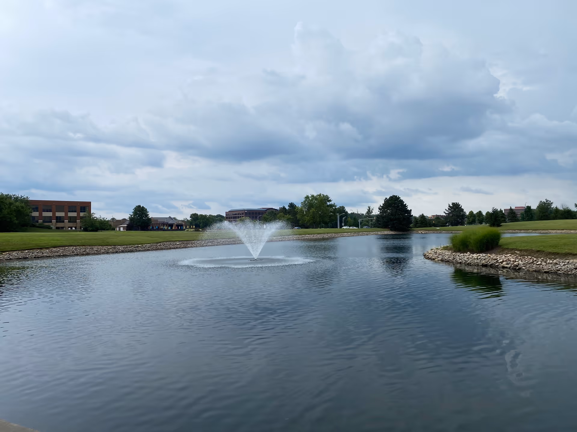 A tranquil pond with a fountain, surrounded by lawns and distant buildings under cloudy sky.