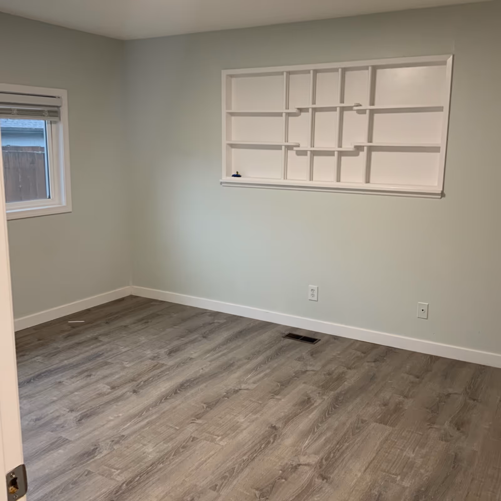 Empty light gray room with a window, wood-like flooring, and a built-in white wall shelf.