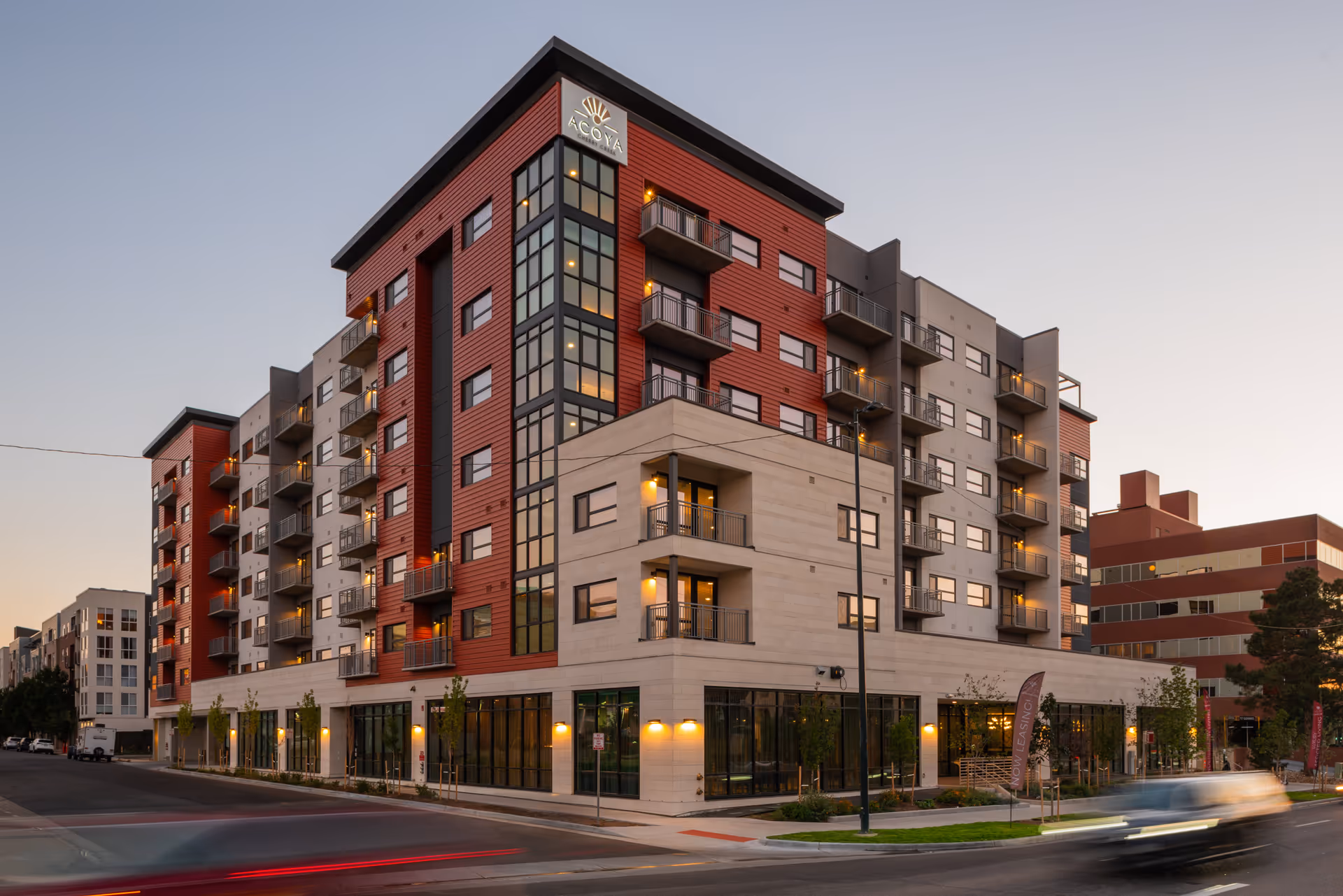 Front exterior of the multi-story Acoya Cherry Creek building at dusk showing balconies, street-level windows, and illuminated signage.