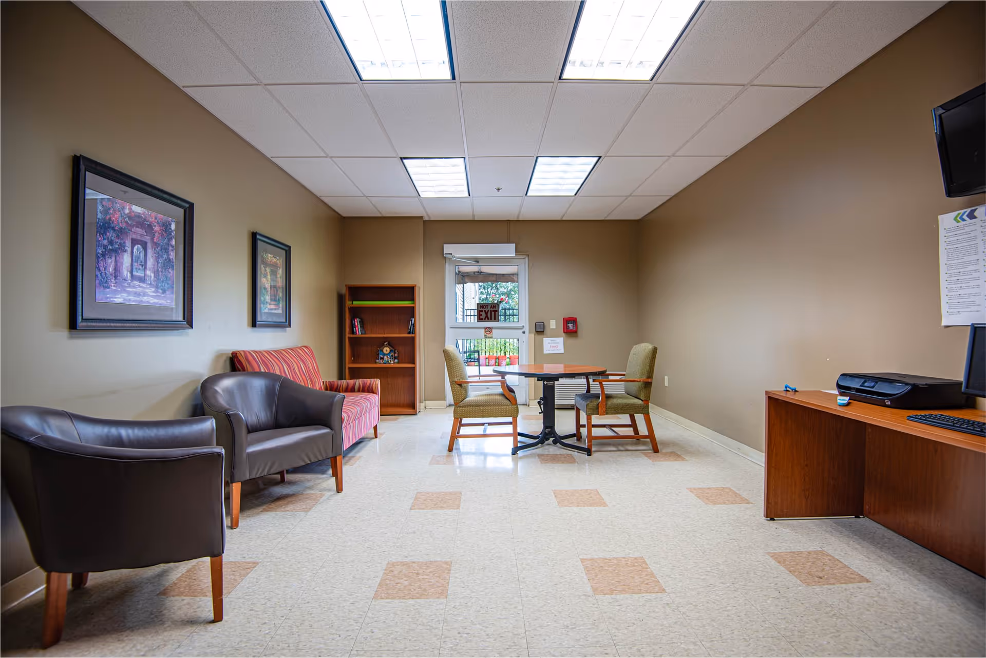 Interior senior living facility lounge with seating, bookshelf, desk, and exit door.