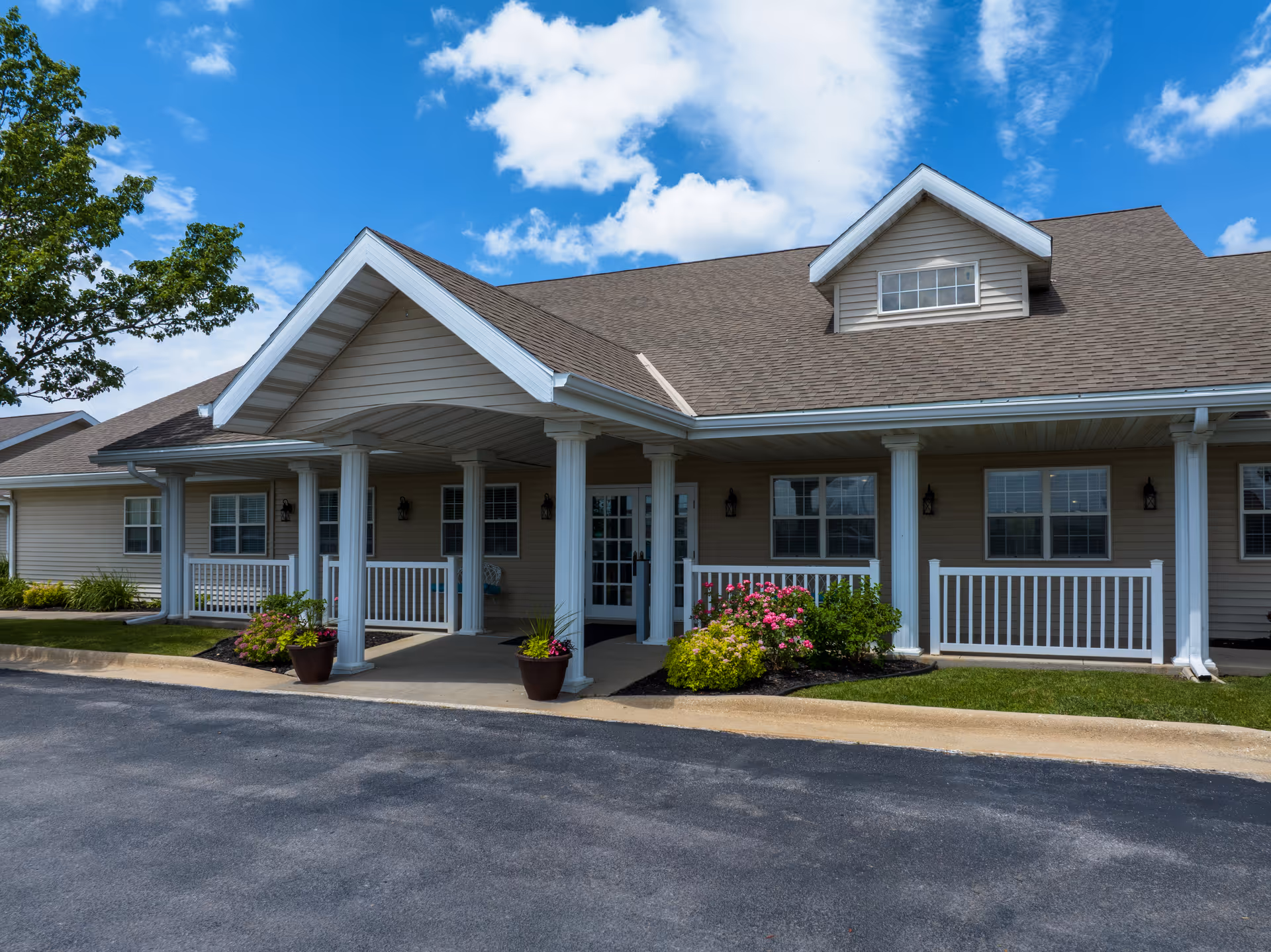 Exterior view of a single-story senior living facility building with beige siding, a covered entrance supported by white columns, a small porch with white railings, potted plants, and a well-maintained garden with flowers under a partly cloudy blue sky.