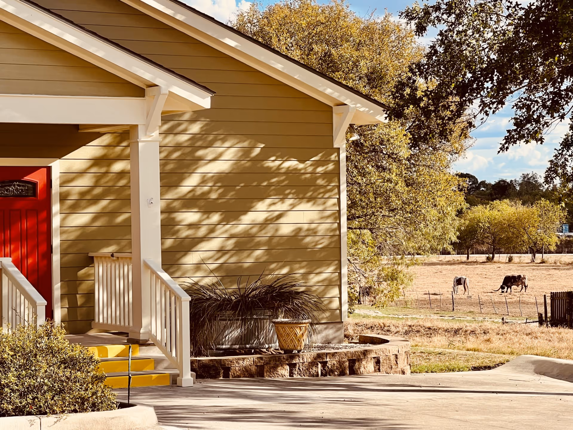 Front view of a beige building with a red door, porch steps, and surrounding farmland.