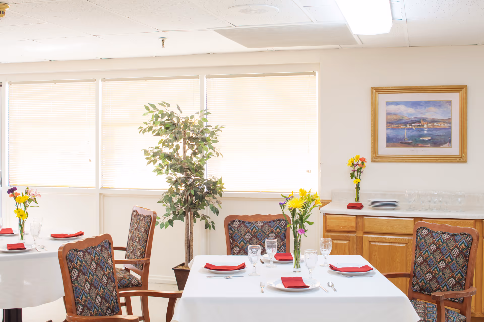 Bright dining room with set tables, upholstered chairs, flower centerpieces, and a sideboard with a framed painting.